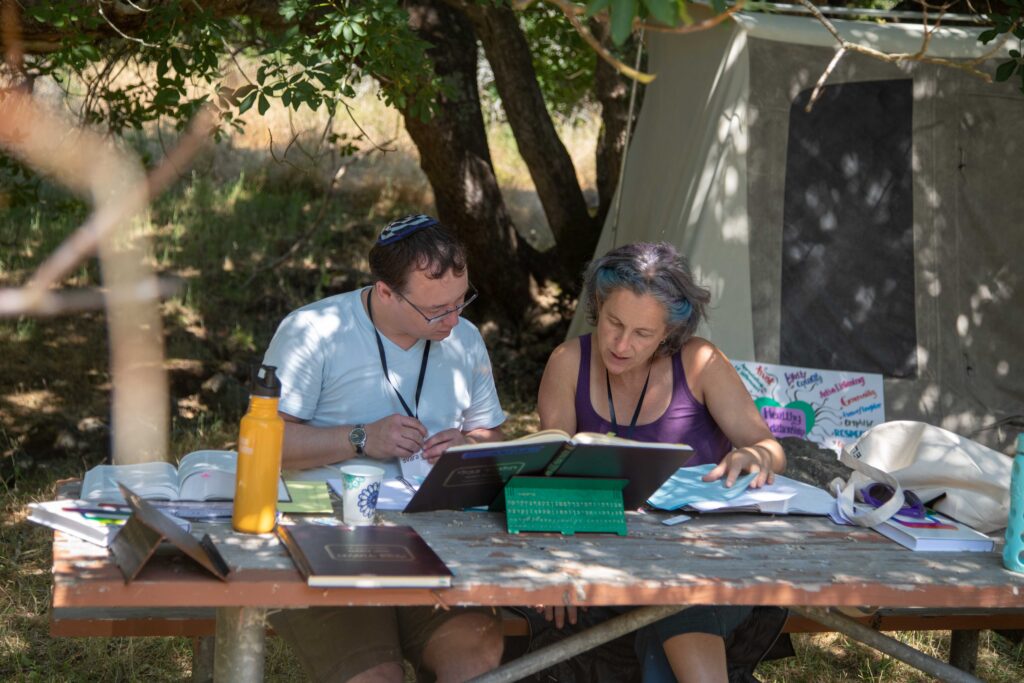 Two people are seated at a picnic table, outside amid grass and trees. The person on the left is wearing a blue kippah atop short brown hair and the person on right has grey and blue hair and is wearing a purple tank top. The table is overflowing with Talmud learning tools like dictionaries, shtenders, and colorful SVARA materials. Behind them is a tent and a colorful sign attached to it.